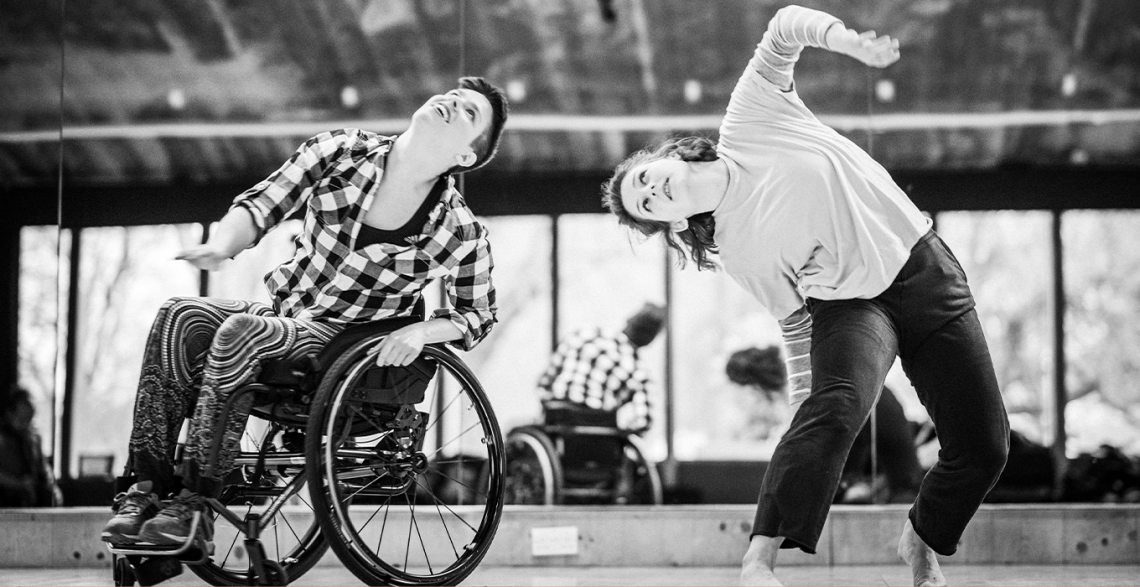 ID: Black-and-white photo of SG2 dancers Kat and Abi moving together in the studio. Kat is white, with short, dark hair. She is wearing a checkered shirt, floaty trousers with a psychedelic pattern and dark trainers. Sitting in her wheelchair, she is leaning to the left, looking up. Abi dances next to Kat on her left side. She is a white, standing dancers with shoulder-length, dark-blond hair. She is wearing a white, baggy t-shirt over a long-sleeved, striped top and dark trousers. She is barefoot. With her knees bent, she is leaning to the right, towards Kat, looking up, with her left arm mid-move upwards as well. Image by Chris Parkes.