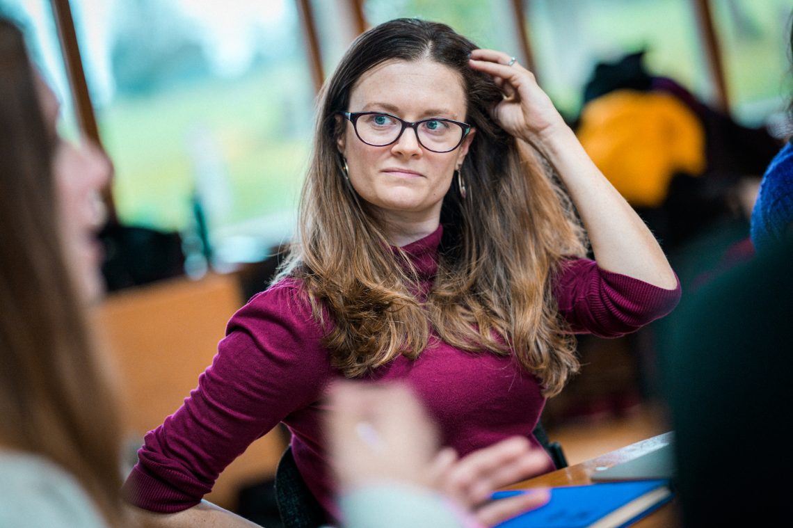 Charlotte sat at a desk listening to someone blurred in the foreground. Charlotte has caramel brown hair draped over her shoulders, blue eyes and she wears glasses. She sits with one hand raised to move her hair way from her face and she is wearing a burgundy top.