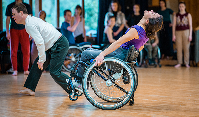 A photo of Laura travelling across a studio, other dancers surround the background of the image. Laura tips backwards in her wheelchair and looks upwards, she is suspended in a wheelie and balancing using her small tail wheel which attaches to the back of her chair. Laura is white, her dark brown hair tied in a bun, she wears a purple top, grey joggers and trainers.