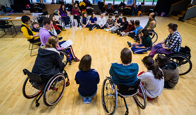 A photograph of the Stopgap team and associate artists, all in a big circle listening and talking to one another. They are in a brightly lit studio space, with wooden panelling, windows behind them open out to a green space. Some people are sat on the floor, others are in their wheelchairs, others sit on chairs.