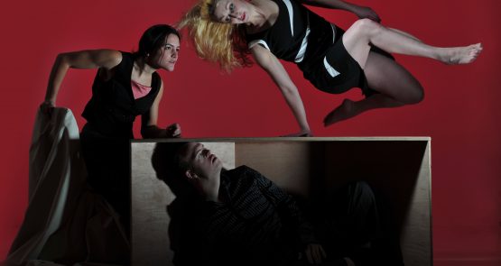 Three dancers, one hides under a table, another leaps over it and the other places a fist on it.