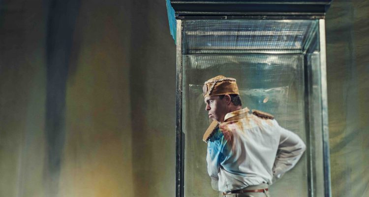 Chris Pavia stands within a clear vitrine, dressed as his dictator character in Artificial Things