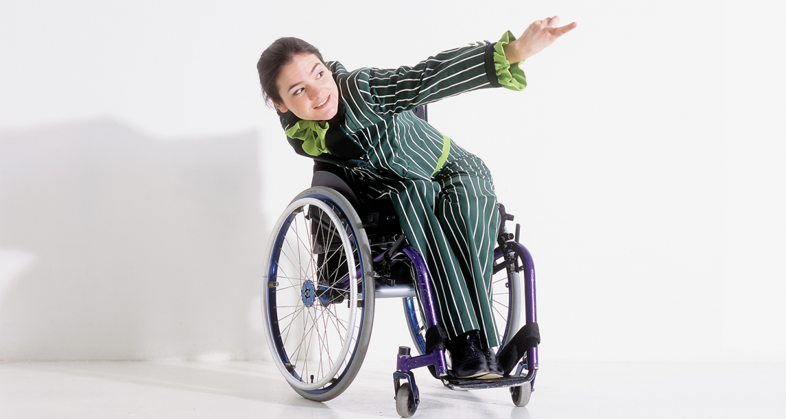 Laura, a wheelchair dancer, in a pinstripe green suit, leans forward and extends an arm.