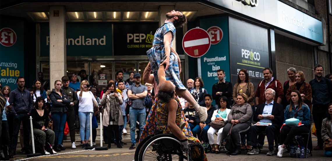 Three dancers during a Frock performance, seated and standing audience watch in front of a storefront on a high-street. 