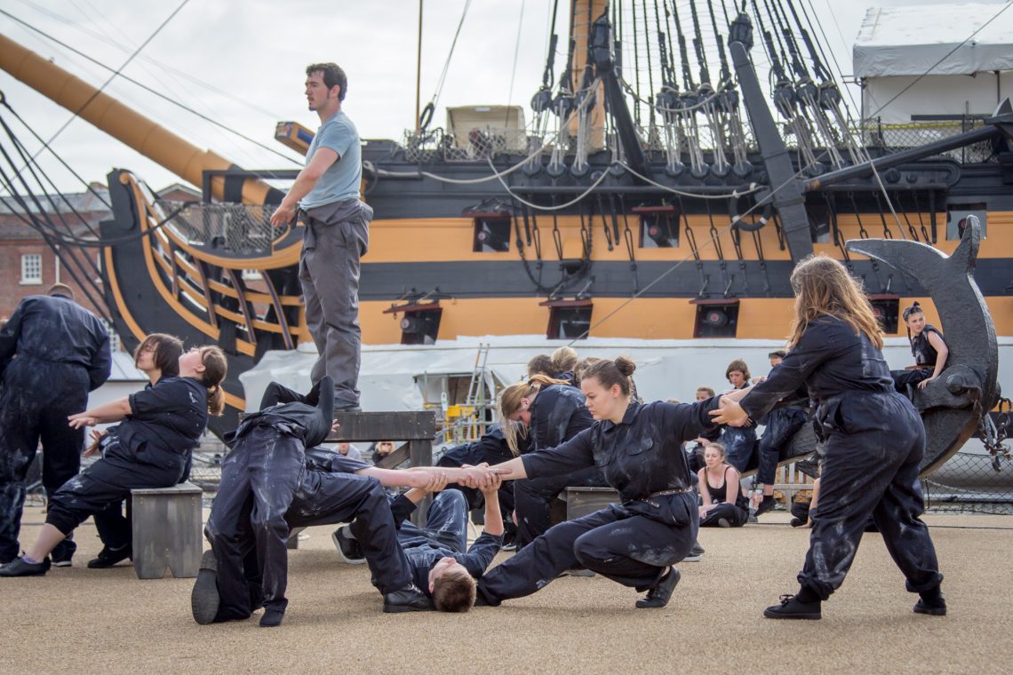 A group of dancers in dark blue overalls reach and pull against each other across the image. Set in front of a wooden ship in black and muddy orange paint.