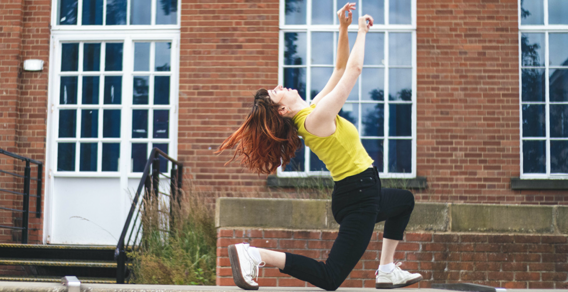 A photo of Lauren dancing outside against a red-brick building with large windows. Lauren lunges forward, her back knee touching concrete floor. She throws her head back, releasing her arms upwards and leaning back with the motion. Lauren is white, her long red tipped hair flows loose as she dances. Lauren is wearing black skinny jeans, a chartreuse yellow vest, and white trainers