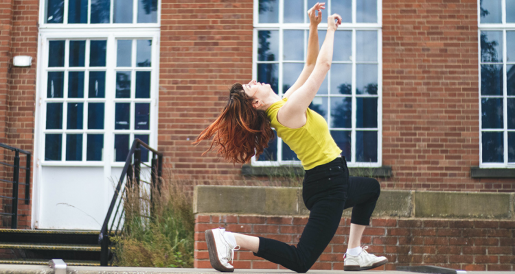 A photo of Lauren dancing outside against a red-brick building with large windows.