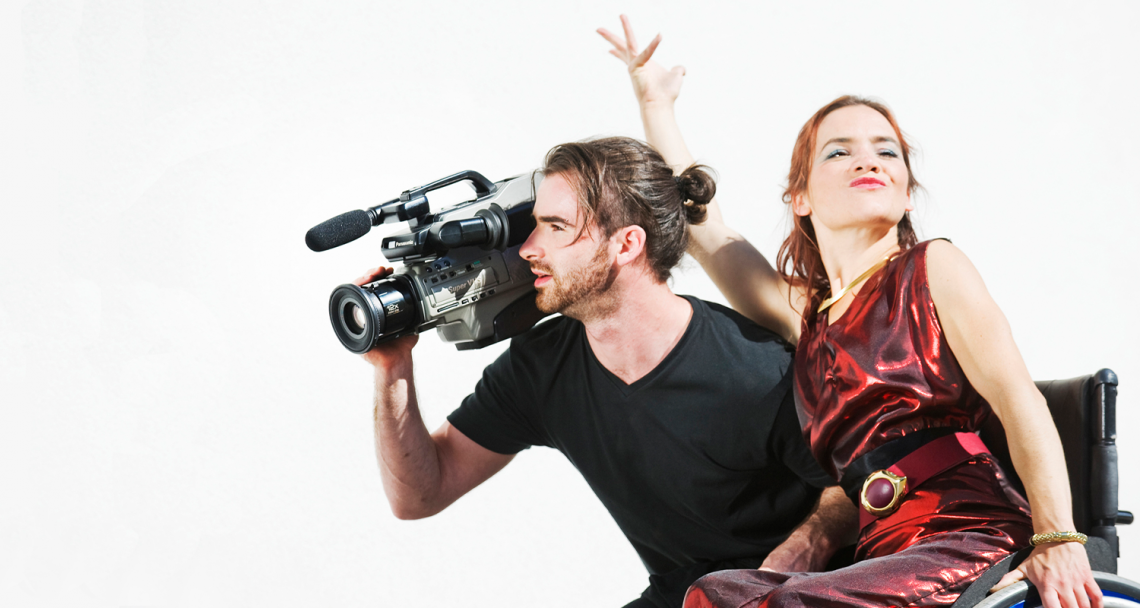 SPUN Productions. Two dancers side by side, one holds a big black film camera and looks through it, the other flamboyantly holds her hand up.