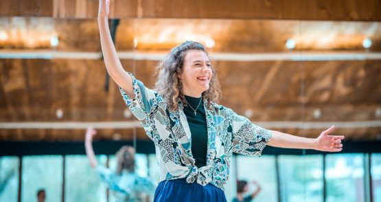 Photo of Millie in the studio, laughing, her arms in a 4th ballet postion (left arm to the side, right arm above her head).