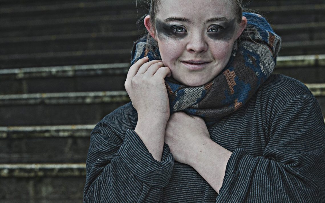 Portrait of Hannah Sampson from The Awakening. The photo is kept in washed-out grey, blue and brown tones. Hannah is looking directly into the camera. Her eyes are painted with very heavy black make-up that stretches out towards her temples like a pair of wings. The rest of her face is very pale and her hair is tied behind her head. She half smiles into the camera. A patterned scarf in blue, beige and grey tones, which she holds with both hands near her right shoulder, is wound around her neck. Hannah is dressed in a long sleeved grey top with narrow, dark grey horizontal stripes. In the background behind her are grey stone steps.