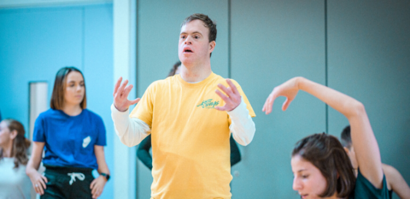 Chris Pavia dancing in a studio with other dancers. he is wearing a yellow t-shirt over a white, long sleeve top. Both his hands are raised to shoulder level in front of him, elbows bent. His facial expression is one of concentration and focus as he looks straight ahead. Photo by Chris Parkes.