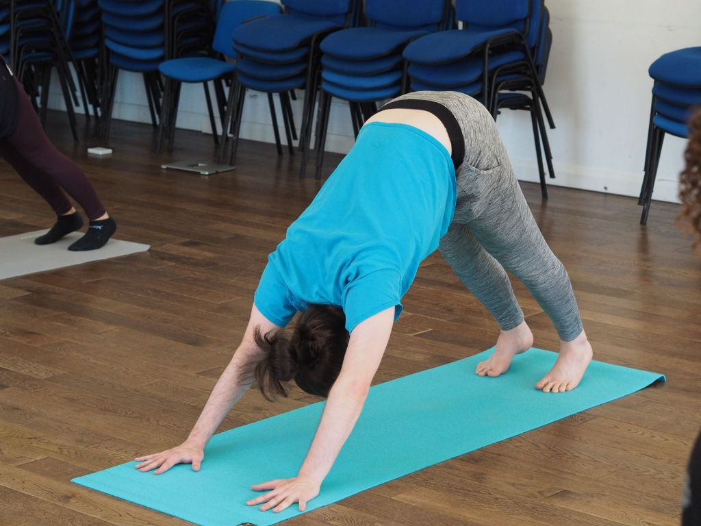 A person in a turquoise t-shirt and grey jogging pants is performing the "downward dog" pose on a turquoise yoga mat.