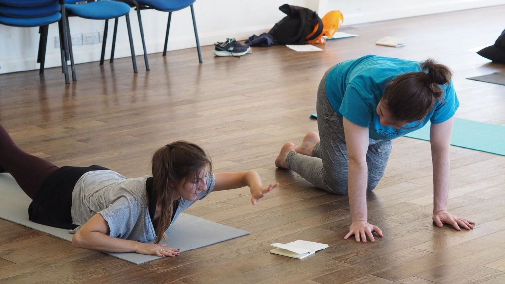 Two young women on the dark wooden floor of a studio. The one on the left is lying on her front on a grey yoga mat, arms bent at the elbows. She is lifting her head, rib cage and left arm off the mat. In front of her is an open notebook. The other is kneeling in the floor beside her. She is on all fours, also looking at the notebook.