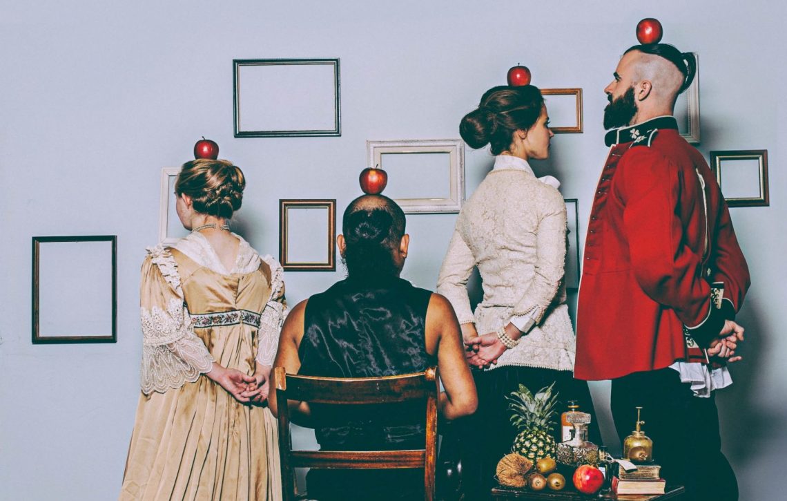 Photo from Stopgap's 2014 production, Exhibition. A group of four dancers are facing a wall hung with empty picture frames, with their backs to the camera. They are dressed in 19th-century style costumes and each balancing a apple on their head. In the foreground a small table with books, bottles, a pineapple, and apple and some kiwi fruit.