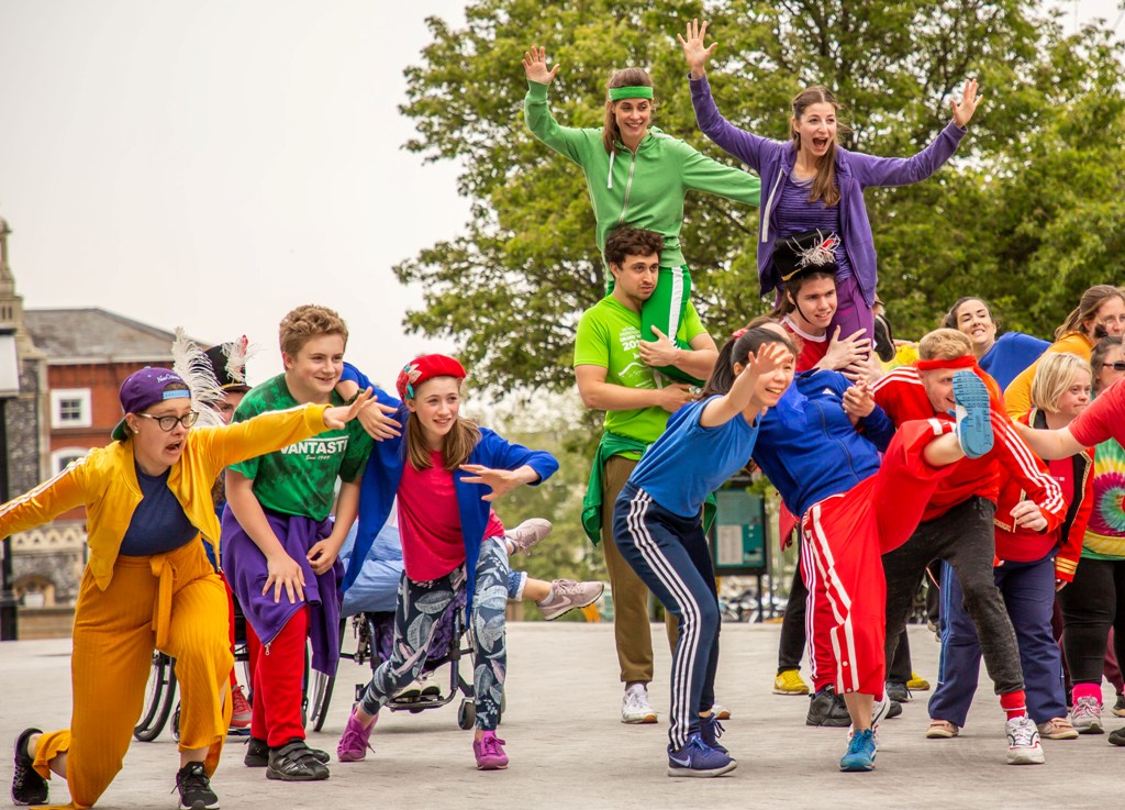 Performance photo of Frippery. A group of young community performers, dressed in colourful tops and trousers, are standing in a row facing the camera, joyfully cheering at something in front of them. Some are throwing their arms forward and one of them is kicking their leg high. Behind them, two male Stopgap dancers carry two of their female colleagues on their shoulders. They are cheering and smiling as well. Photo by Mary Doggett.
