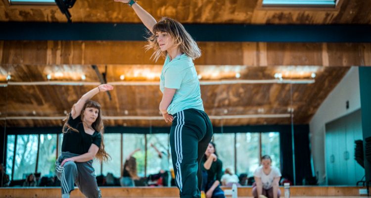 Photo of two female standing dancers with long hair, wearing t-shirts and jogging bootoms with side stripes dancing in the studio.