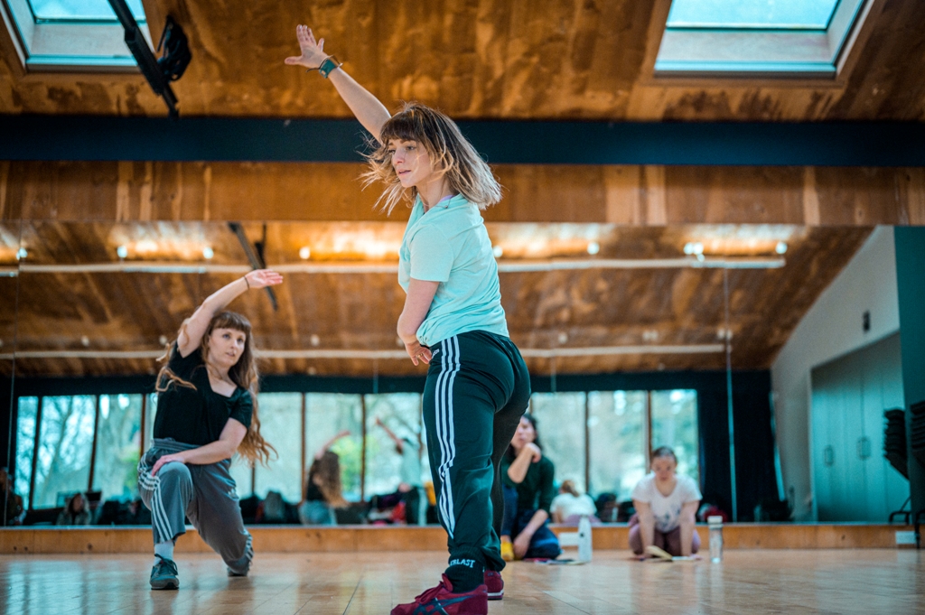 Photo of two female standing dancers with long hair, wearing t-shirts and jogging bottoms with side stripes dancing in the studio. They both put their weight onto the right leg, with knee bent, the other leg outstretched. The dancer on the left in the picture is nearer to the floor, her knee bent in a right angle; the other dancer is standing. Both are mid-move with their right arms stretched long overhead and their left arms across their body. Lucy and Hannah are sitting on the floor, observing. Behind them, all are reflected in the studio mirror. Photo by Chris Parkes.