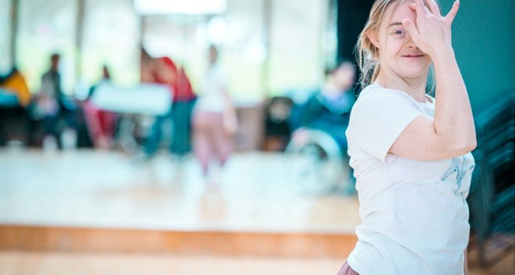 A photo of Hannah, a learning disabled professional dance artist, smiling at the camera. She is in the far right of the image and it cuts off above her waist. Hannah has one hand raised to her face; covering half with one finger resting gently on her forehead. The background is blurred but it's a bright studio space.