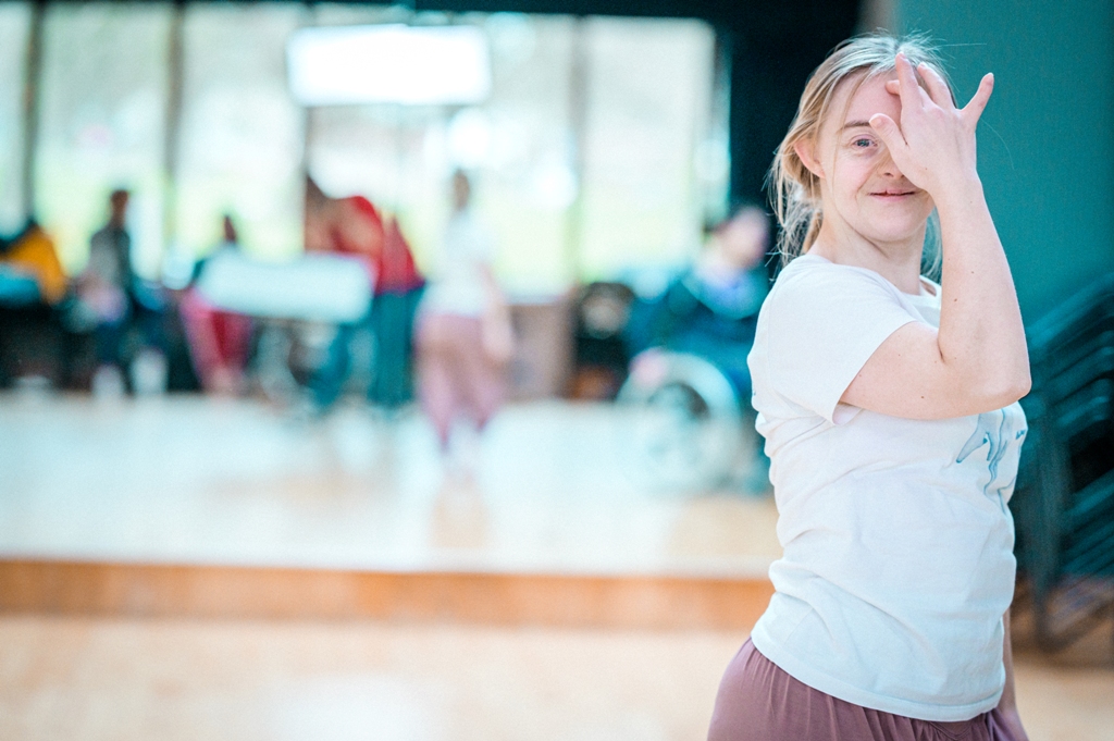 A photo of Hannah, a learning disabled professional dance artist, smiling at the camera. She is in the far right of the image and it cuts off above her waist. Hannah has one hand raised to her face; covering half with one finger resting gently on her forehead. The background is blurred but it's a bright studio space.