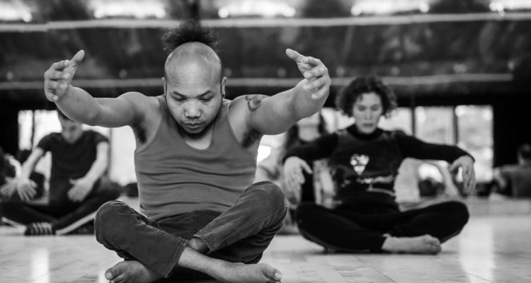 Black and white photo of Nadenh and Amy, two members of the Frock cast, sitting cross-legged on the floor, contracting their upper bodies with arms reaching forward.