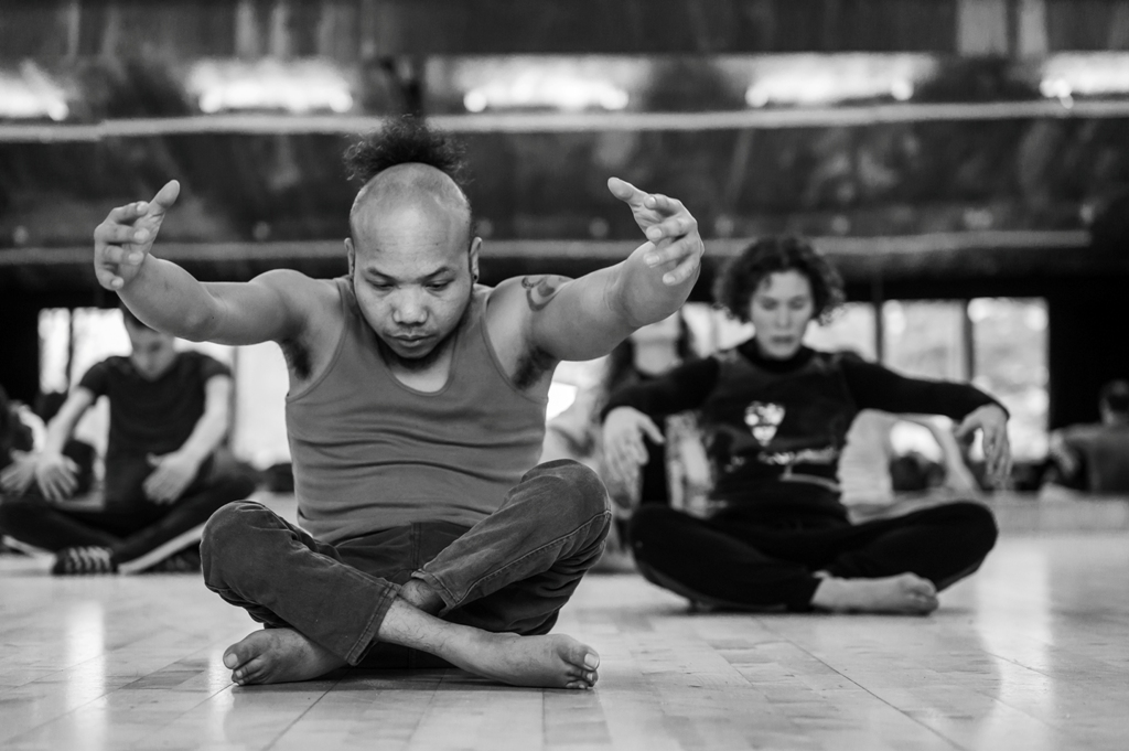 Black and white photo of Nadenh and Amy, two members of the Frock cast, sitting cross-legged on the wooden floor of the dance studio, contracting their upper bodies with arms reaching forward and gaze directed to the floor. The focus is on Nadenh, Amy and other dancers in the background are blurred. Photo by Chris Parkes.