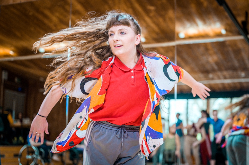 Alice Shepperson dancing in the studio. She is wearing a colourful, floaty, short-sleeved open shirt over a red polo shirt and grey trousers. She pushes her arms back and her long, wavy, light brown hair is being whipped around by her movement. Photo by Chris Parkes.