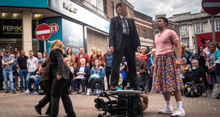 Outdoor performance of Frock, In the centre, Nadenh is lying on his side in his wheelchair. KJ is standing on the top wheel, wearing a black suit, white shirt and tie. To their left, two female dancers in black suits and ties are twisting around each other. To their left, Christian stands and looks at KJ, wearing a pink t-shirt and floral skirt.