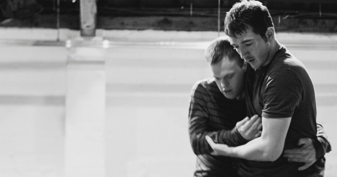 Black and white photo of male dancers Chris Pavia and David Willdridge in close embrace. They are side by side on the right side of the photo, one arm around each others' waist, the other arm held in front of their bodies; David's left hand gently cradling Chris's right elbow. Both are looking down, to a point on the floor in front of them.