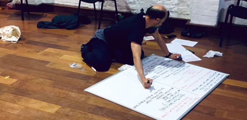 A photo of Tom Goodwin, kneeling on a wooden floor writing on a big whiteboard.
