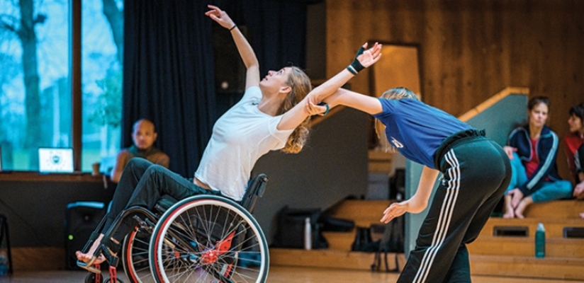 A duet of a wheelchair and a standing dancer in the studio. The wheelchair dancer is wearing a white t-shirt and black trousers. She is tipping back in her wheelchair with the front wheels lifting off the ground. She has her left arm stretched upwards. her right arm is moving upwards in a curve and she follows it with her gaze. The standing dancer is dressed in a blue t-shirt and black trousers with white stripes at the sides. They are bending forwards away from and with their back to the camera, holding the wheelchair dancer's left arm with their left hand. Both their upper arms are directly connected to their hands.