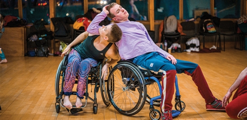 A duet of a female and a male wheelchair dancer leaning into each other. The male dancer is leaning casually back in his wheelchair, dressed in a lavender top, blue-red trousers and trainers. He is leaning with his right elbow on the female dancer's shoulders, his head tipped sideways into his hand. The female dancer is twisting around his upper arm, looking upwards to his hand. She is wearing a teal tank top and colourful patterned trousers. She is barefoot.