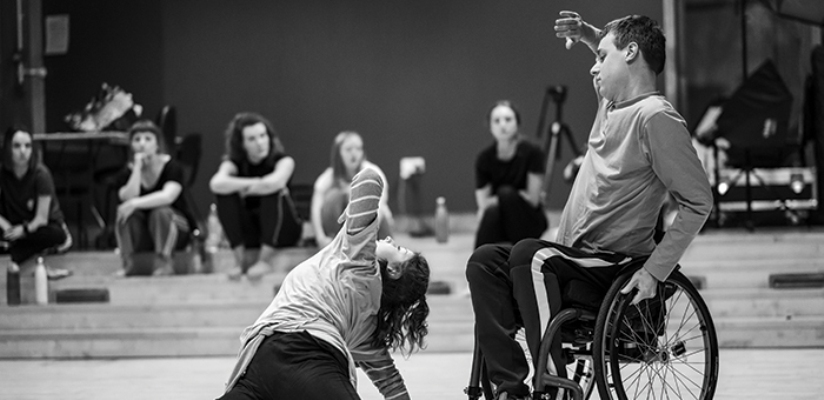 Black and white photo of a duet of a male wheelchair dancer and a female standing dancer. The wheelchair dancer is dressed in a long-sleeve top and dark trousers with white side stripes. he is holding on to the left wheel of his chair with his left hand. Hid right hand is lifted in front of his face, palm outwards. The female dancer is on the floor before him, only partly visible. She is wearing a white t-shirt over a striped top. She is leaning on her right arm, her back to the male dancer, and is twisting her body to the left, with her left elbow leading the movement. She is looking up, her dark hair floating over her shoulders.
