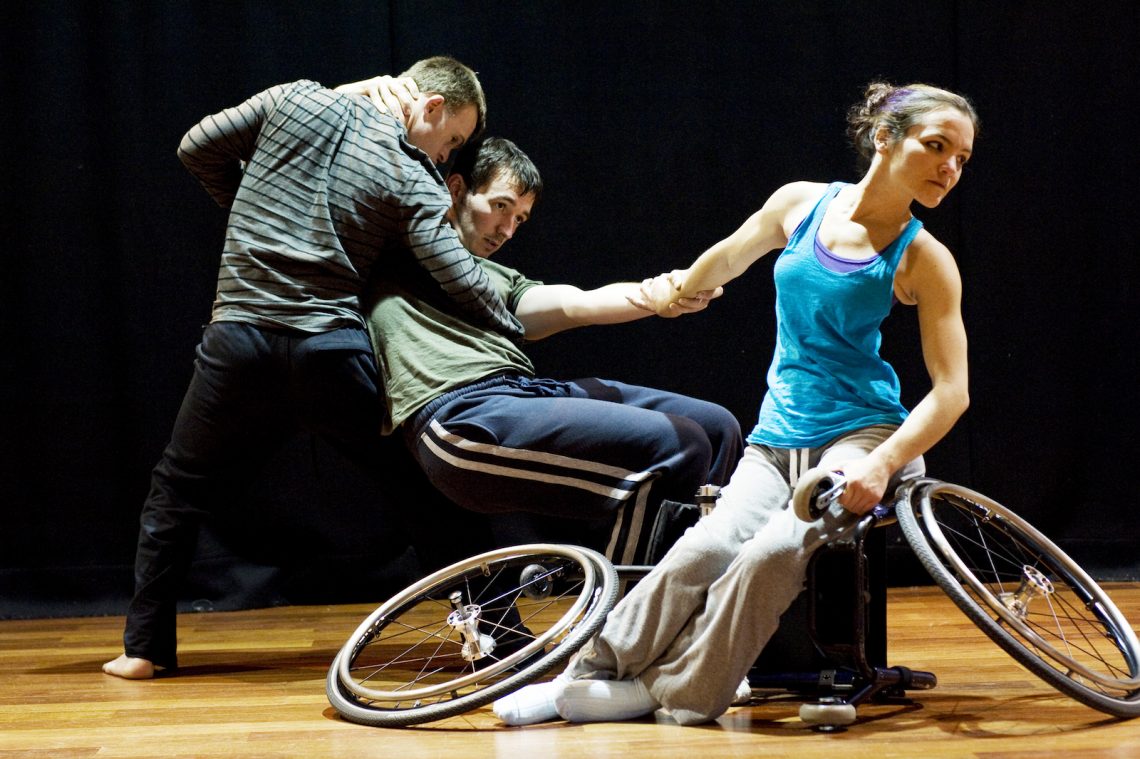 Laura, Chris and David in rehearsal. Laura sits on a dismantled wheelchair. Chris supports David as he sits without a chair between them.
