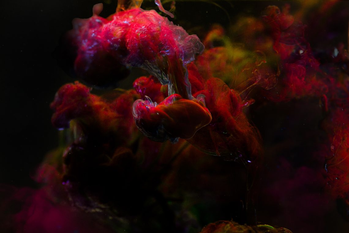 An abstract textural photo of pink and red plumes of swirling liquid spreading through dark water. Creating a space nebula of deep red clouds and hazy greens, highlighted by bright electric blue and white static ripples.