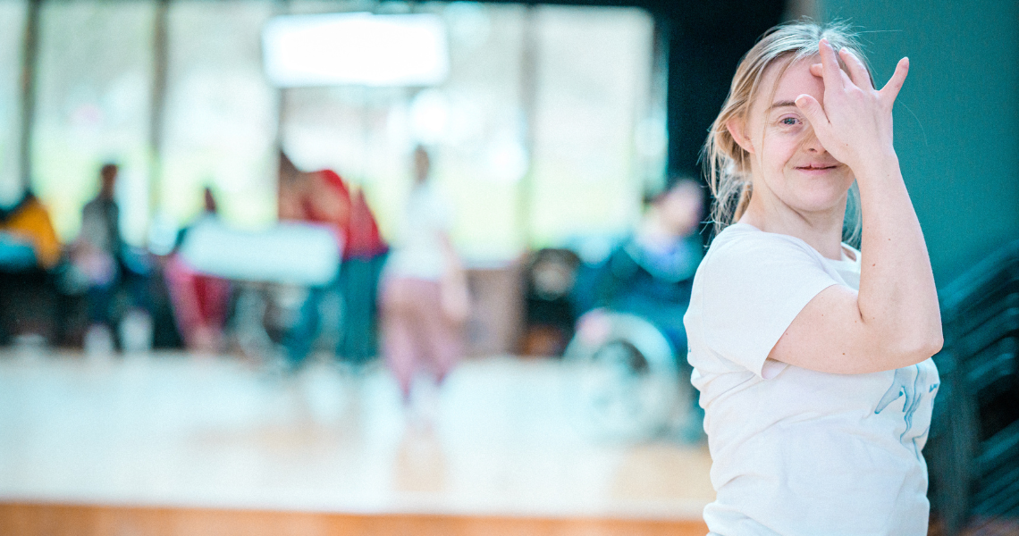 A photo of Hannah Sampson, a white learning disabled person who has Down syndrome. Hannah has blonde hair tied back from her smiling face, she touches a fingertip to her forehead and gazes at the camera with blue eyes. Other dancers are reflected in the mirrors in the background out of focus. 