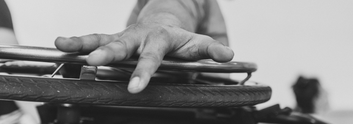 Black and white photo of a hand over the horizontal wheel of a wheelchair. 
