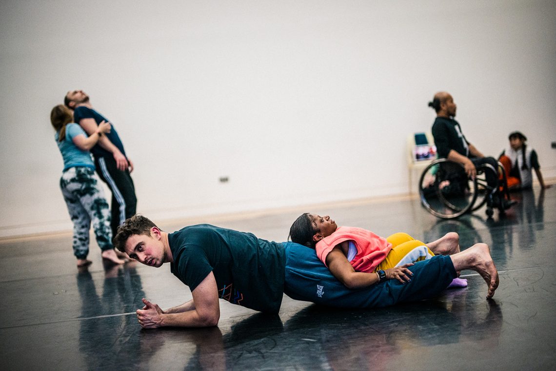 Six dancers in a bright studio. In the foreground Christian, a non-disabled white male dancer with shot dark hair, pauses in a plank position, hands clasped. On his legs rests Mo, a short-statured Disabled Black woman with dark hair tied back, she reclines back and grips Christian's calves. in the background to either side, two other duets are blurred.