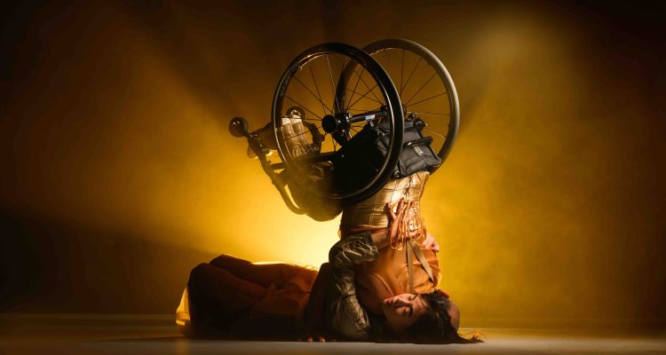 Dancers Emily and Nadenh during a duet. Emily, a non-disabled east asian woman lays on her back and calmly embraces Nadenh, a wheelchair dancer, lifting him and supporting his back as he balances in a headstand. The wheels of his lightweight black manual chair catch focus as he suspends his lower body and inverts. There is a sense of serenity and tension, Emily’s face is calm as they embrace but Nadenh’s face is hidden, and the strength of the balance is immense. Golden light spills from behind them, catching on the rich browns and ambers of their costumes. Photography by Chris Parkes.