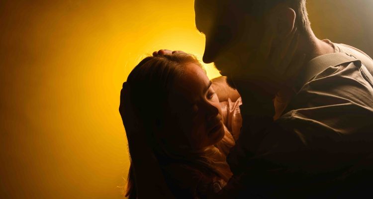 An intimate close up of dancers Hannah and Jannick embracing, filling the right two thirds of the frame. Faces close and shadowed almost in silhouette, they grasp each other's cheeks and hold one anothers heads gently. A halo of golden back light glows around their faces. Photography by Chris Parkes