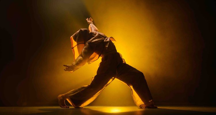 Christian, a white non-disabled standing dancer, in a wide lunge, silhouetted as he drapes his shirt over his face. He is leaning backwards, his torso going back over his hips, his left leg bent in front of him and the other foot behind in a gentle arch. His arms are raised stretched out beside his body.