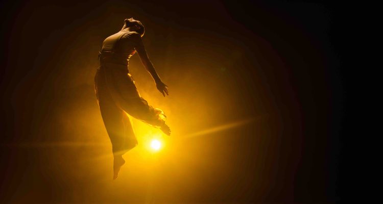 Jannick, a white standing dancer, caught mid-jump, his upper body arching as if pulled upwards by his chest. A strong golden light in the middle of the image cuts through haze and the dark background.