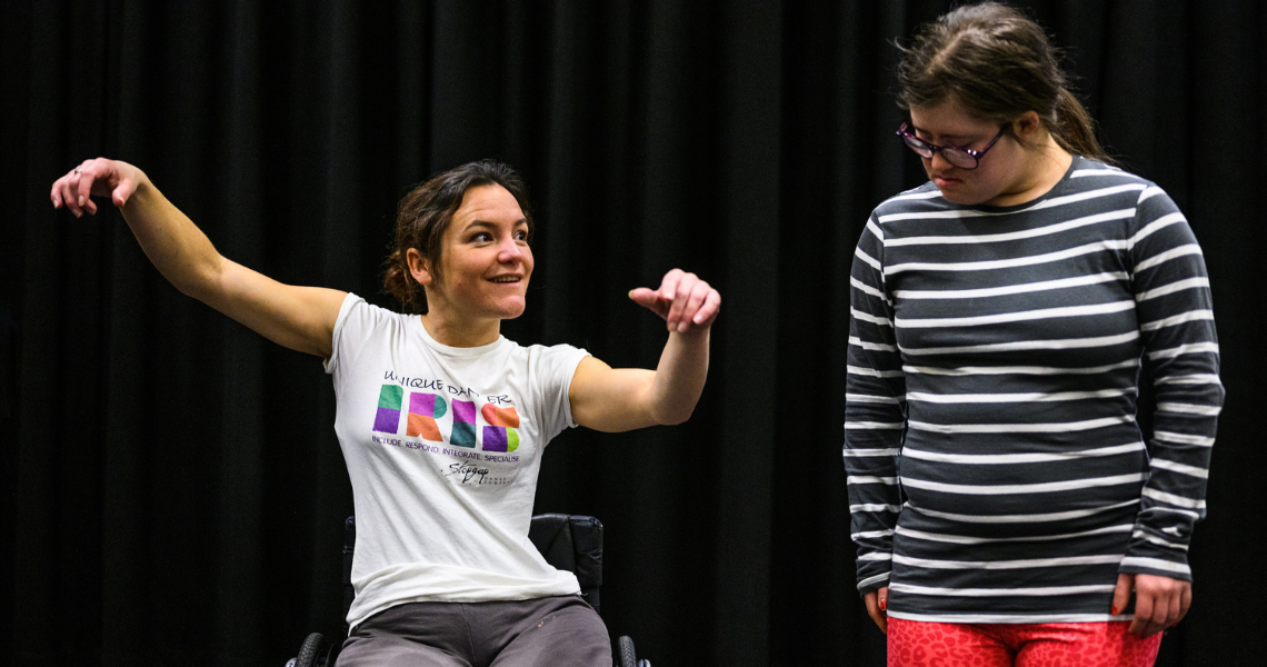 Co-Artistic Director Laura teaches some young dancers, she is sitting in her manual wheelchair smiling at the dancer on the right. Laura's arms float up to the side. On the right a young dancer watches Laura's movement.
