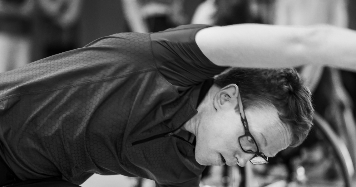 A black and white image of Dermot during class, his body horizontal as he leans over and reaches his right arm overhead, he looks down to the floor in concentration. 