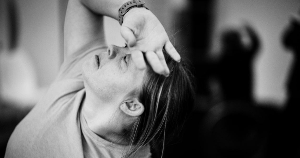 A close up black and white image of Hannah, a white learning Disabled person with Down syndrome. Hannah raises her gaze upward and arches back as they rest the back of their hand on their forehead, elbow high behind them.