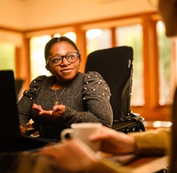 A black woman in a power wheelchair sits at a table and talks to someone out of frame.